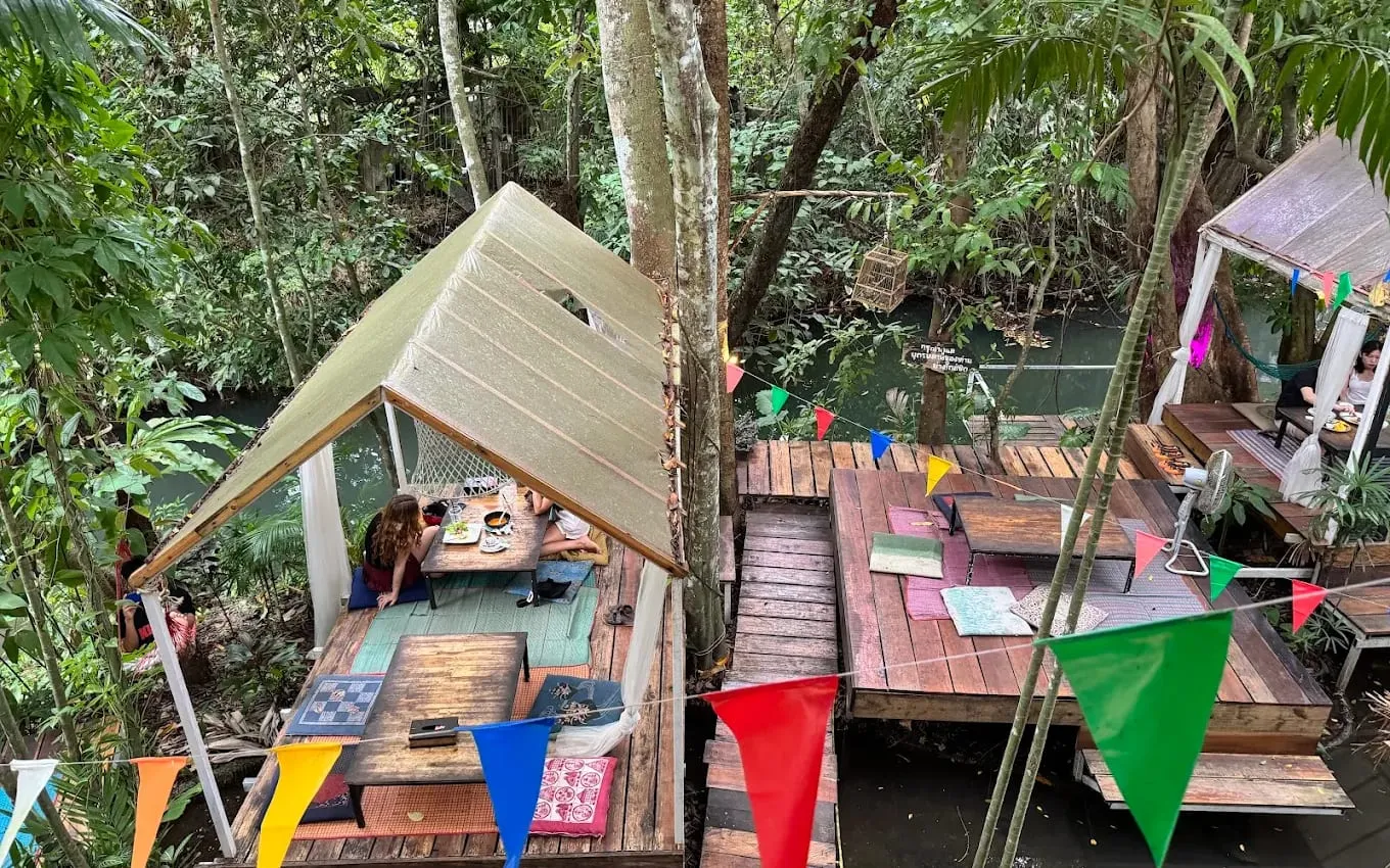 Riverside seating at Baan Ton Mai seen from above: low tables on wooden decks over a jungle river, one under a pitched canopy where two guests sit on floor cushions, with strings of coloured flags across the front
