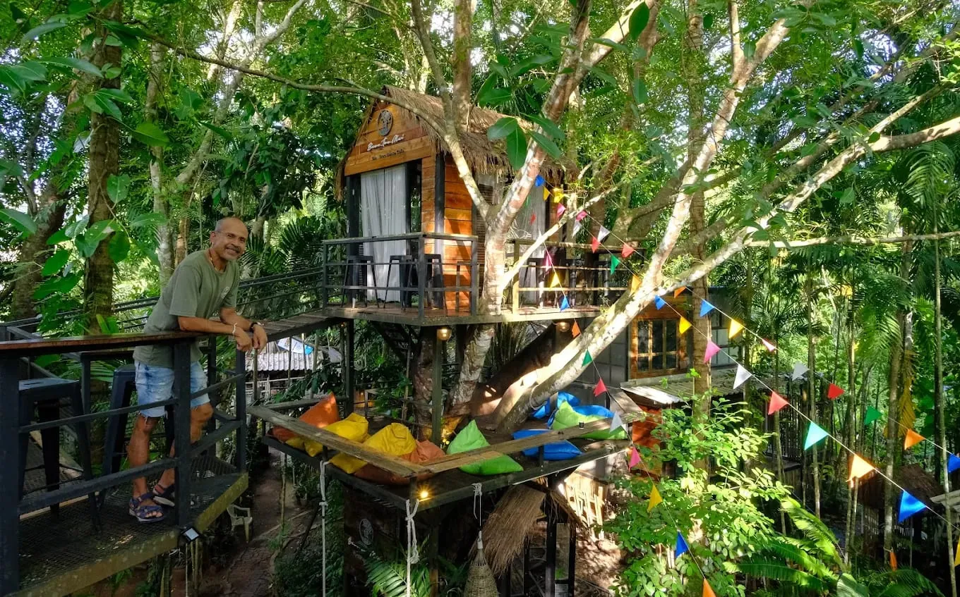 A man leans on the railing of a raised timber walkway at Baan Ton Mai, beside a wooden treehouse cabin built around a tree trunk. Below the cabin, a deck holds bright floor cushions, and strings of coloured flags run through the surrounding jungle.