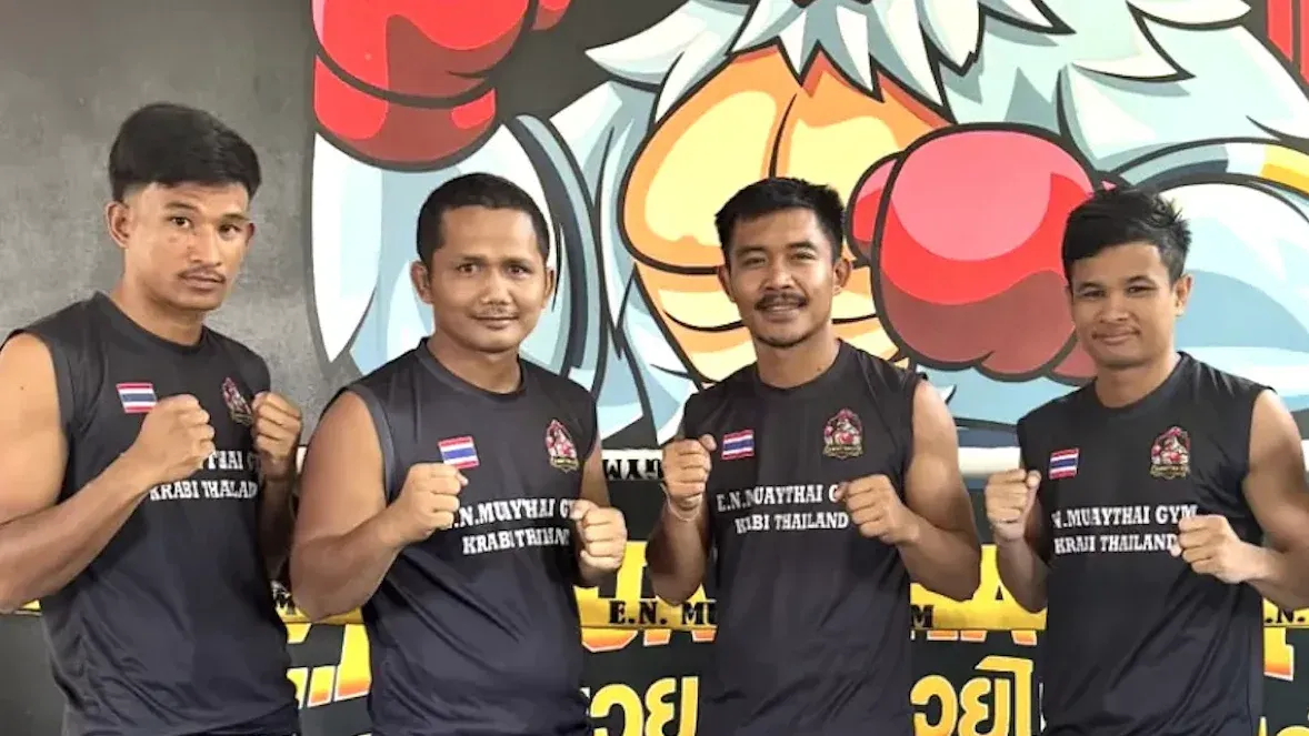 Four EN Muay Thai coaches in black sleeveless E.N. Muay Thai Gym shirts, fists raised, in front of a colourful painted mural with red boxing gloves