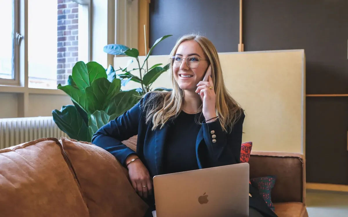 A smiling woman on a phone call, sitting on a sofa with a laptop on her lap